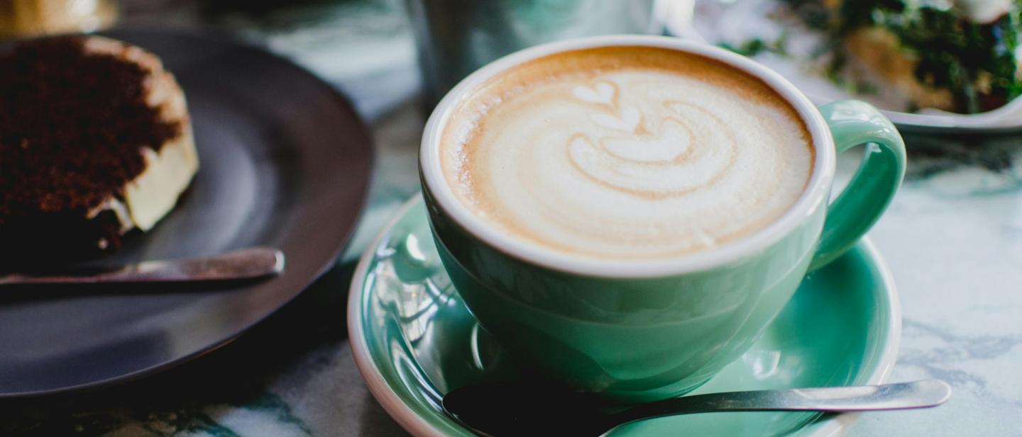 Teal cup and saucer with a latte coffee and some cakes in the background